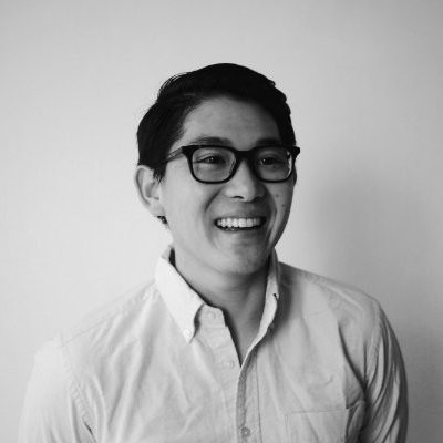 A dark-haired man with black-rimmed glasses smiles while wearing a white buttondown shirt