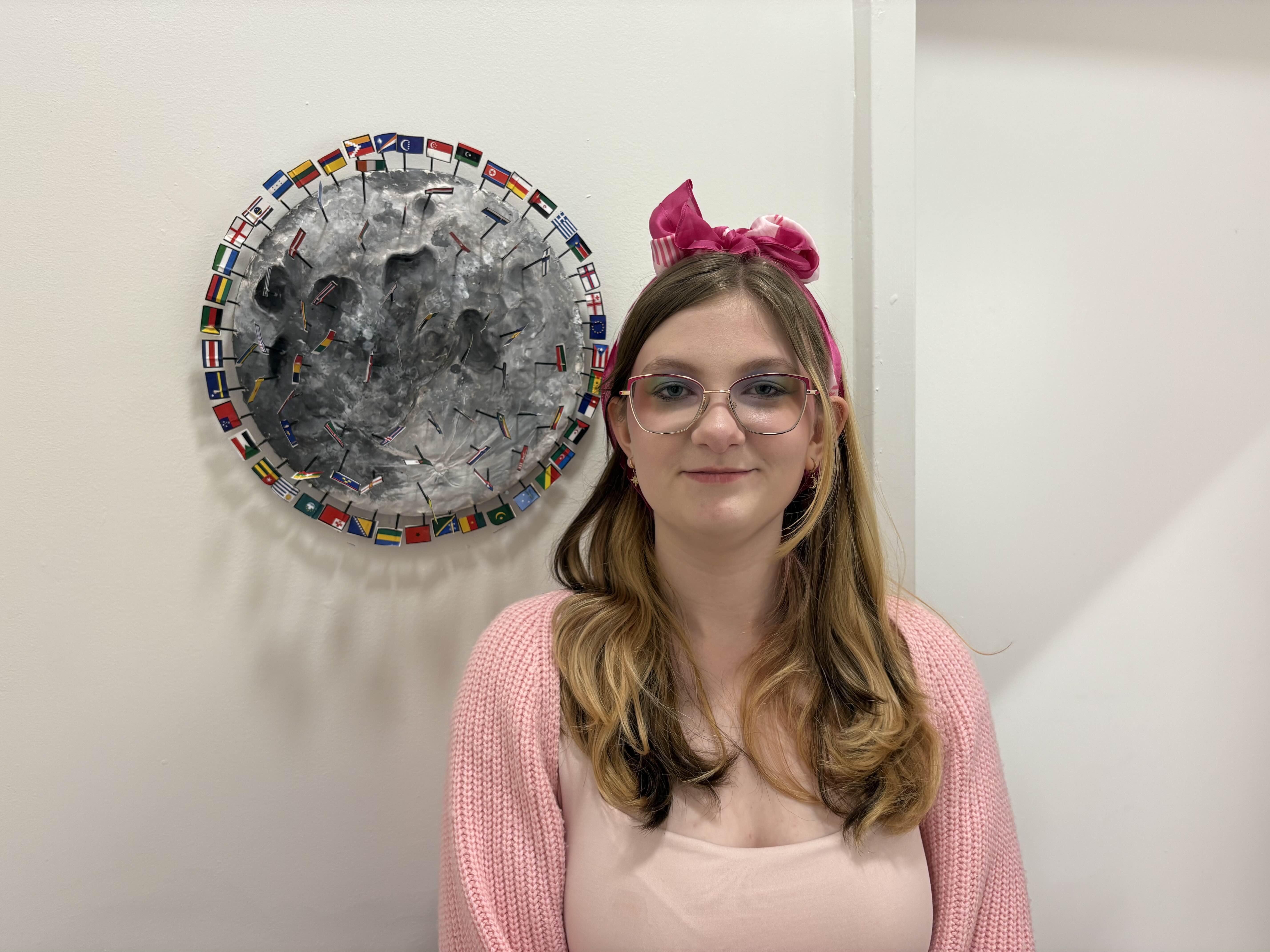 Student standing next to a wall-mounted moon sculpture with small flags surrounding it.