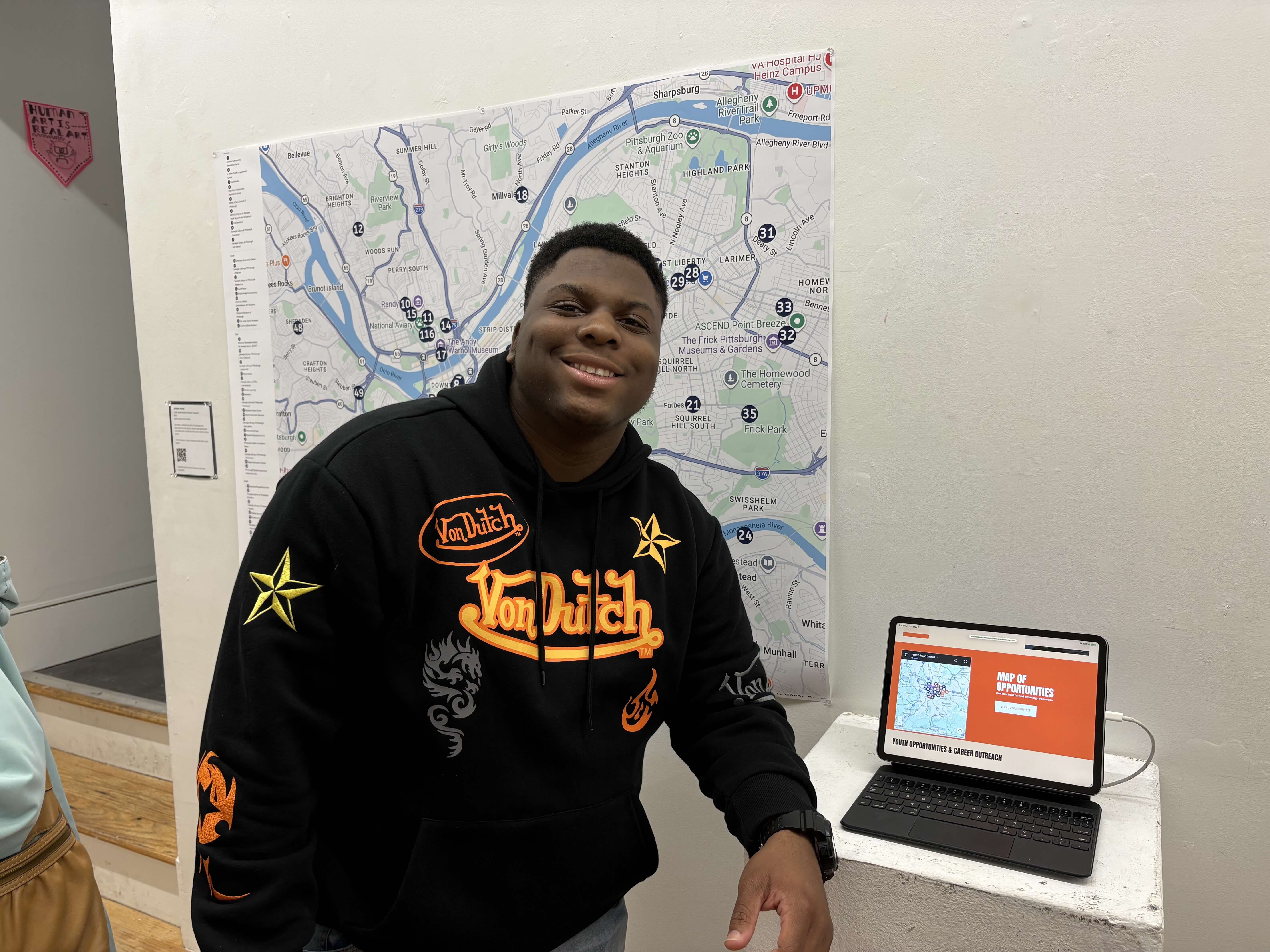 Student smiling in front of a large printed map of Pittsburgh and next to an iPad displaying a website called YOCO.