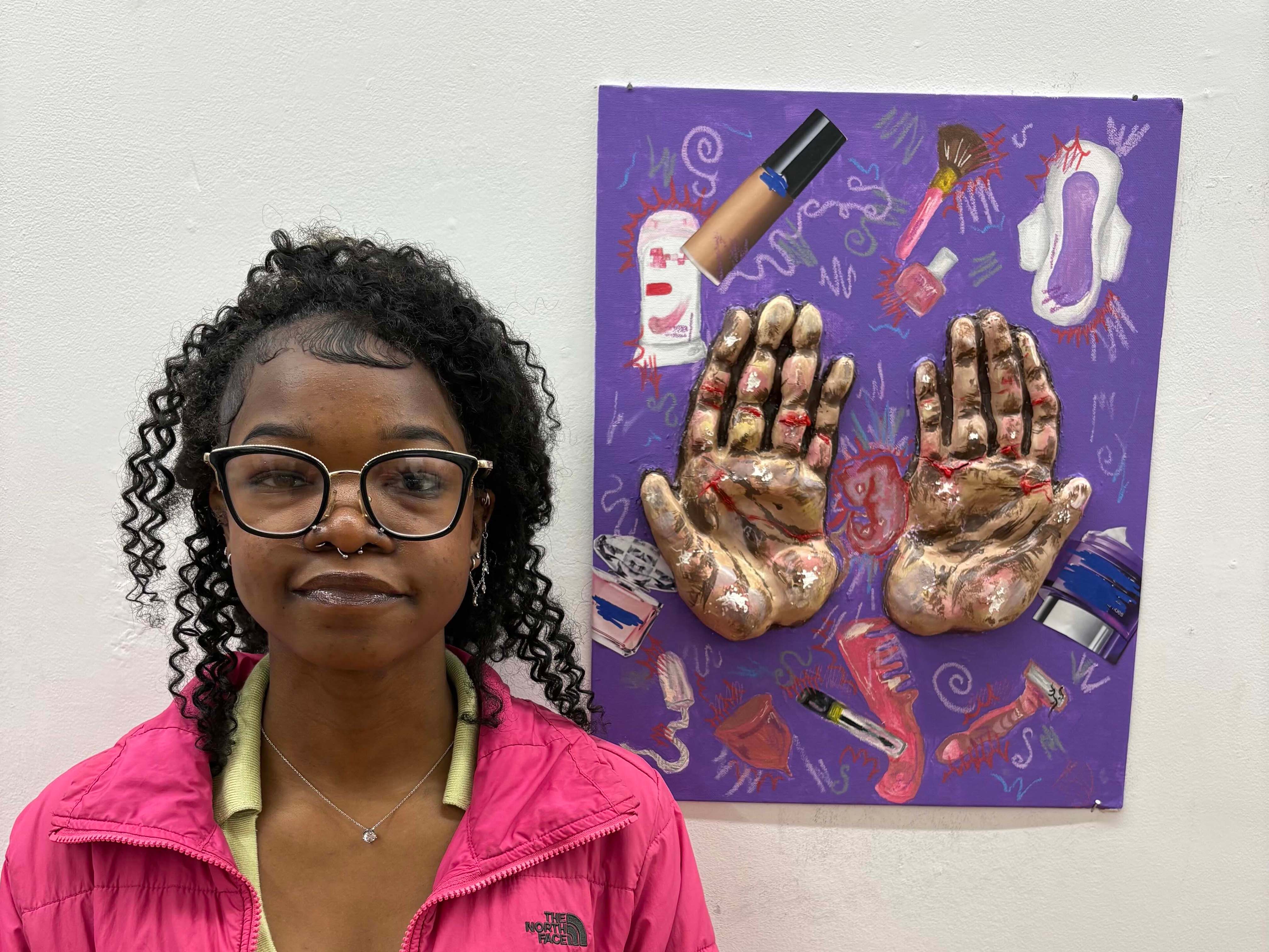 Student standing next to painting with sculpted, three-dimensional hands attached to it.