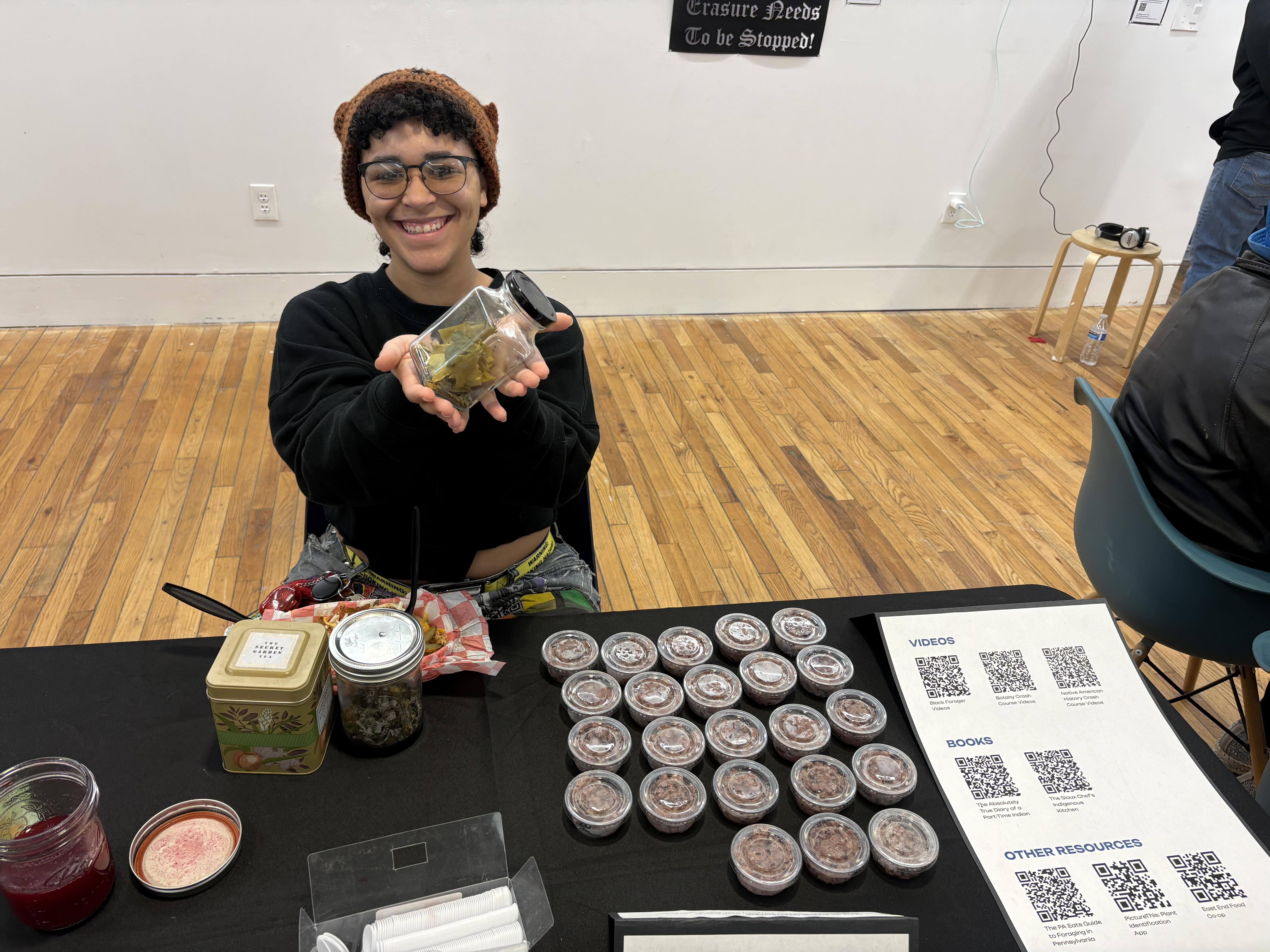 Student holding a jar of foraged food, sitting behind a table with small cups of desserts.