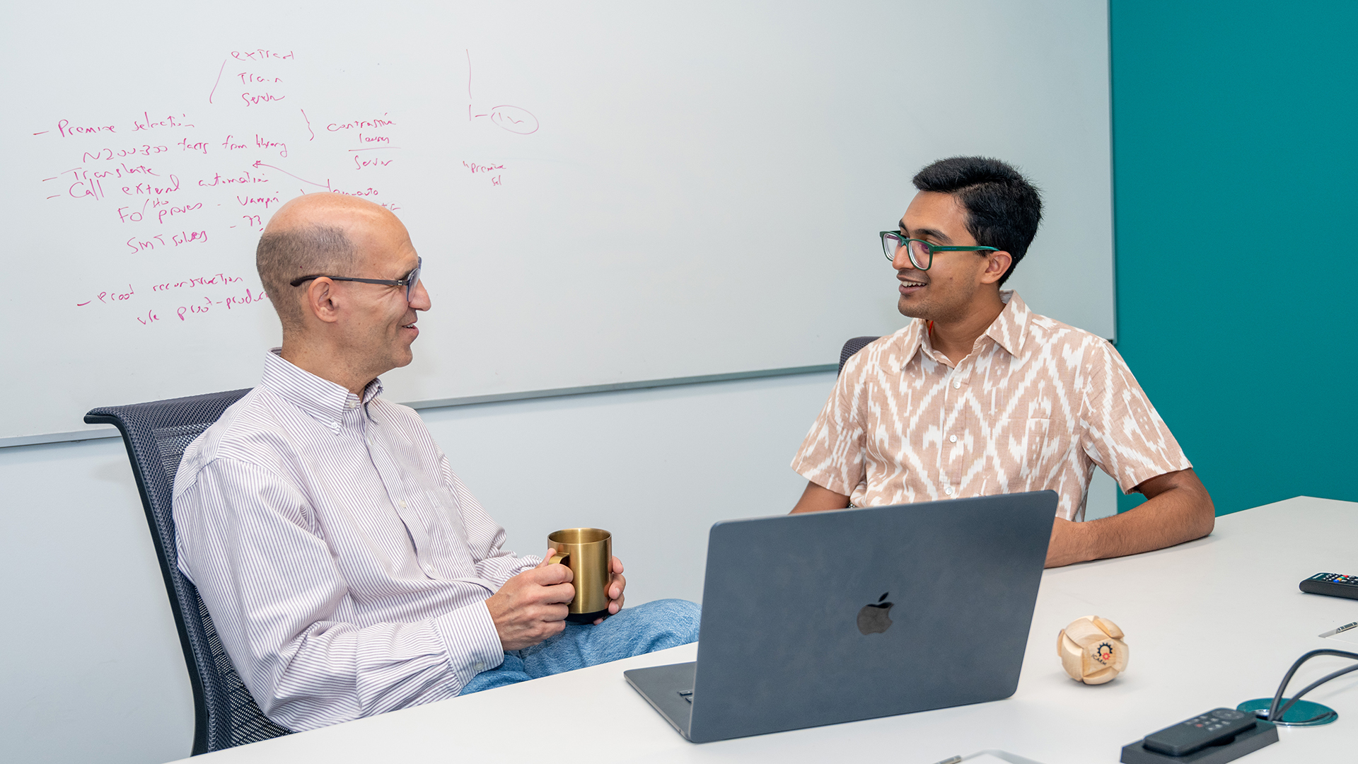 Jeremy Avigad and Sidharth Hariharan sit at a table together. They both smile as they talk. In the background, there is a whiteboard with mathematics equations on it.