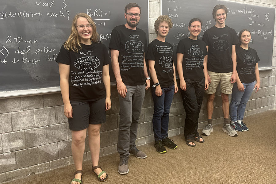 Six people stand in a row wearing similar mathematical phrases on their shirts