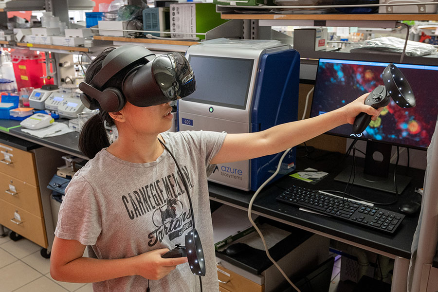 A woman wearing a virtual reality headset moves her arms while a monitor shows the interior of an infected cell that she is navigating