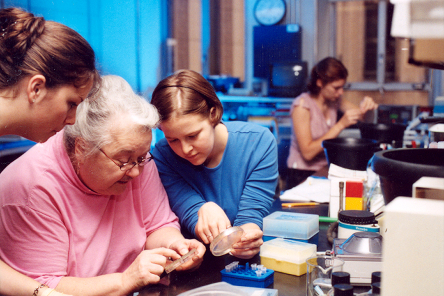 An elderly female teacher in a lab, guiding three female students as they conduct an experiment.