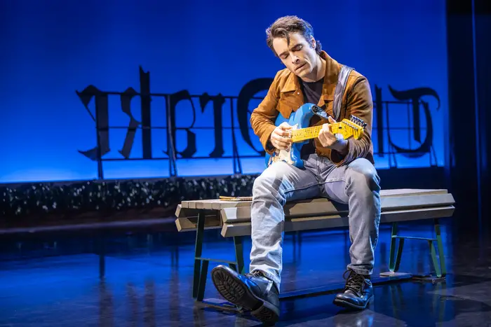 Man wearing jeans and a brown leather jacket sings and plays a blue and white electric guitar on a stage set to look like a rooftop.
