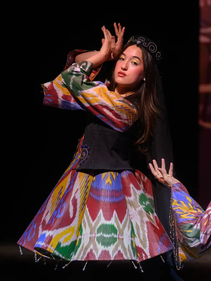 A woman wearing a delicate sparkly crown stands while leaning back slightly and holding her hands in a fluttering motion near her face. She is wearing a multicolored dress with a pattern of thin lines shaped in an abstract pattern made up of purple, yellow, red, green, and beige. She is also wearing a dark denim vest over the top part of the dress.