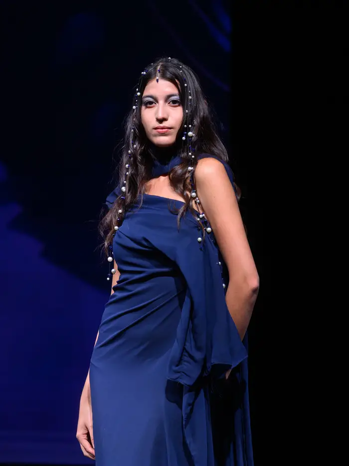 Woman poses with one shoulder forward while wearing a navy blue one-shoulder dress with delicate ruching towards the right side of her waist. In contrast with the deep blue of her dress, she has pearls woven into her long dark hair that frames her face.