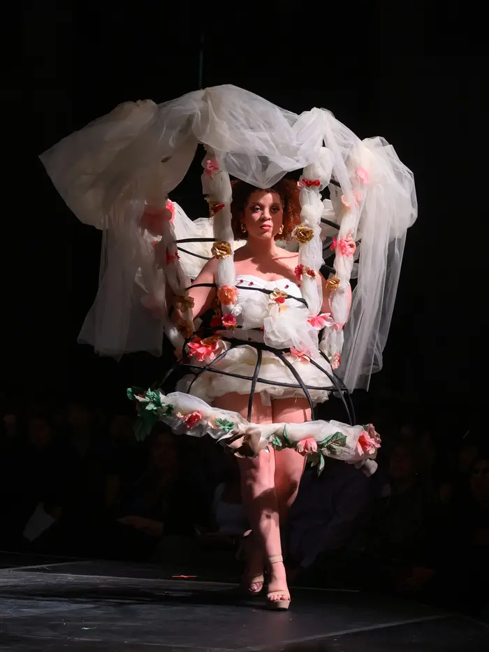 A woman walks down the runway, wearing a cage in the shape of a hoop skirt, with the metal exposed and intertwined with white fluffy tulle fabric and pink and green flowers. She has an inverted version of the hoop skirt starting at her waist and extending outward as it comes up around her head. The top part is also intertwined with tulle and flowers, and has a sort of veil draping down from it to frame the model.