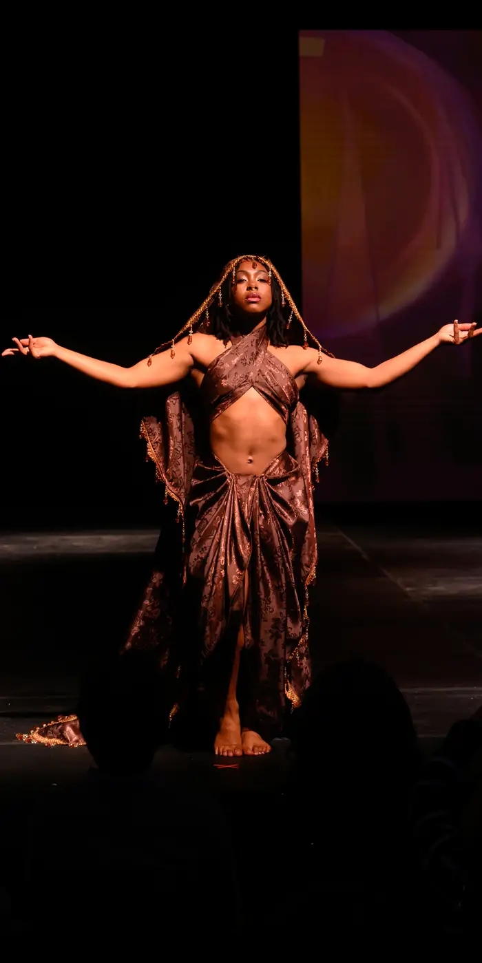 A woman stands with her arms outstretched to the side, wearing a pleated skirt, wrap bra top, and head covering - all made of the same brown brocade material. The head covering has strategically placed jewels dripping down over her hair, face, and arms.