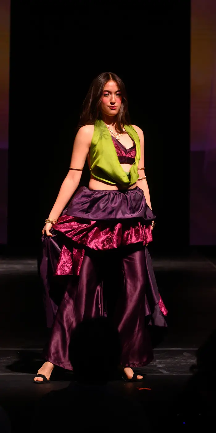 Woman stands holding out a flared skirt that is made up of multiple purple, pink, and wine-colored layers. The skirt is paired with a pink bra and a chartreuse green vest overtop.