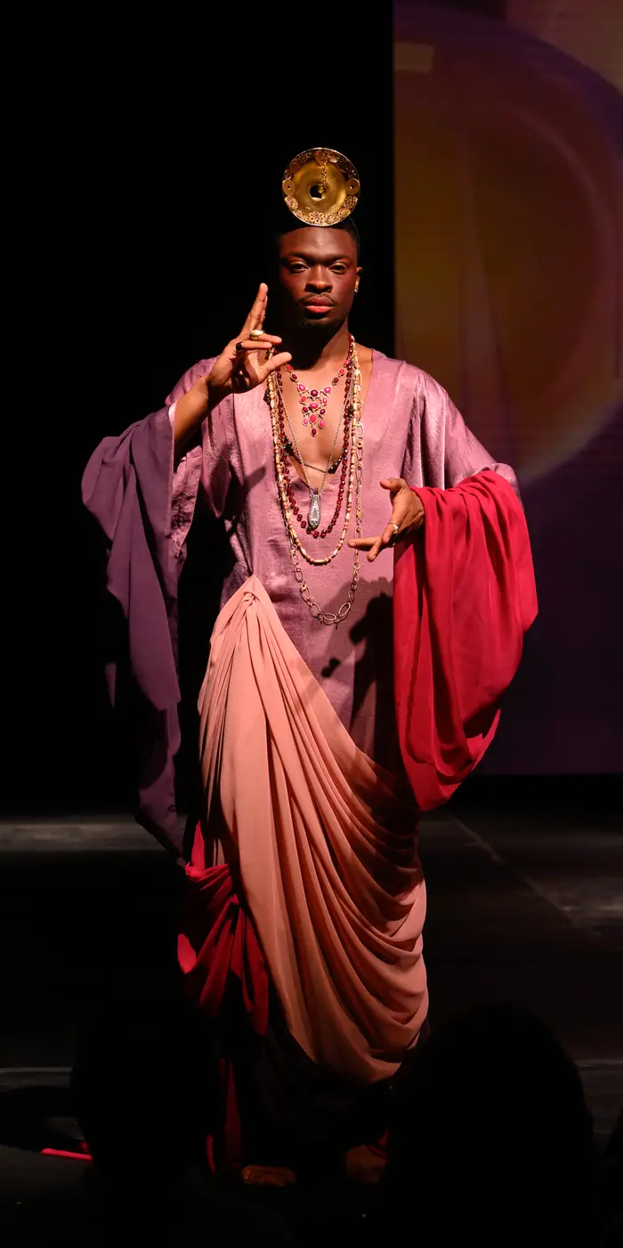 A man poses holding one hand out slightly in front of him, with his other hand tilted up with two fingers pointing up just in front of his face. He is wearing a gold circle attached to a headdress and has multiple layers of long gold and jeweled necklaces. His outfit is a series of gorgeously draped fabrics, with one pale purple shirt that stretches to his legs, and a pale pink fabric draped starting at his waist and going around his body. His arms are draped in elegant sleeves, one in red and one in purple