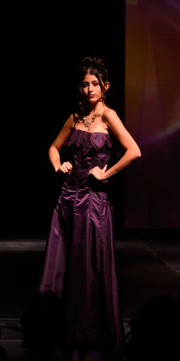 Woman stands with her hands on her hips, with one shoulder forward. She is wearing a strapless floor-length plum purple gown with delicate fuchsia piping along the bust. She is also wearing a necklace of three silver flowers.