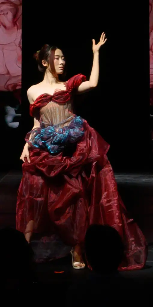 A woman poses with her face tilted to the side, holding her arm up and out. She is wearing a majestic gown made of a layered red tulle skirt, a gorgeous iridescent corset-like top with red tulle around the bust extending into small sleeves around her upper arms. The dress also has a thin ruffle of iridescent blue fabric gathered around her waist, adding contrast and drama to the voluminous burgundy skirt.