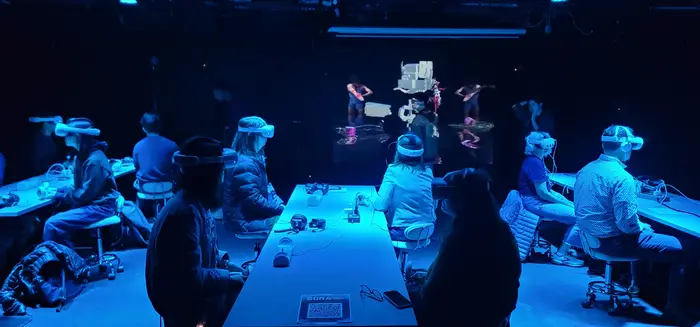 A group of people sits at tables while watching a dance story unfold on VR headsets