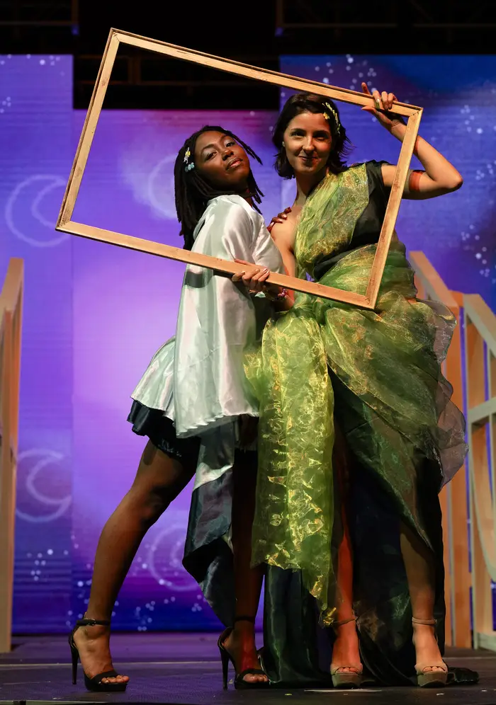Two models hold a rectangular gold frame around their faces, with one wearing a short shimmering green-silver dress with long sleeves, and the other wearing a one-shoulder midi-length gown of shimmering yellow-green fabric with a wide organza waist that tapers as it cascades down her legs