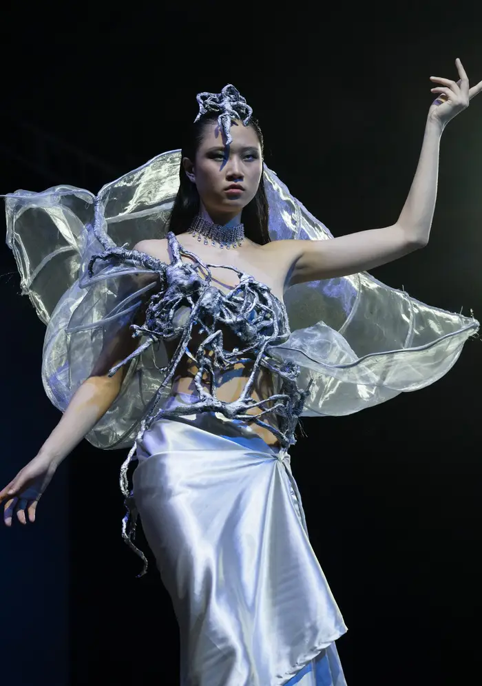 Model wearing a metallic silver asymmetrical pencil skirt, with a top made of silver wiring around her chest that has sheet shimmering metallic fabric undulating around her back. She is also wearing a thick silver shoker necklace and a silver wire headpiece