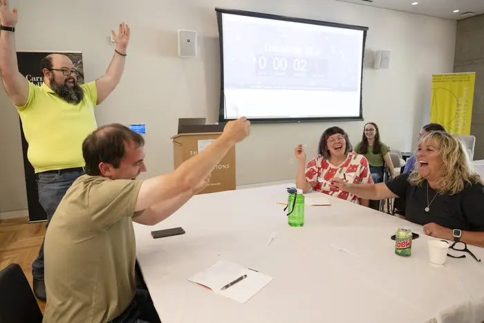 A man in a yellow shirt raises his arms in victory standing behind a group of three people, a man and two women, seated at a white table, who also react to sunglasses, a green water bottle, and plastic utensils in the center of the table.