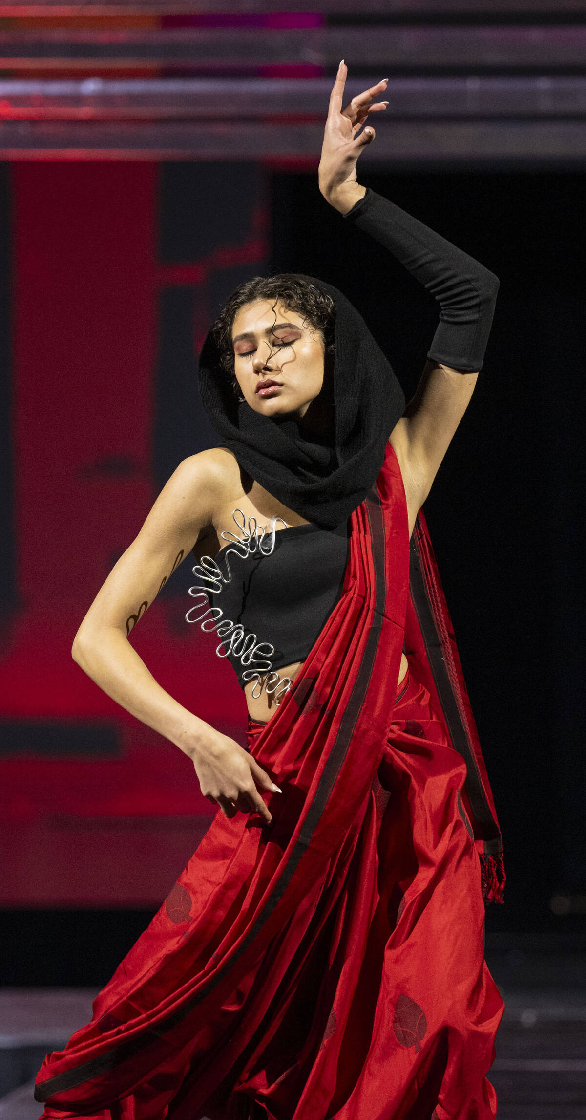 Model wearing black top and silk red pleated garment with curved wire accents.