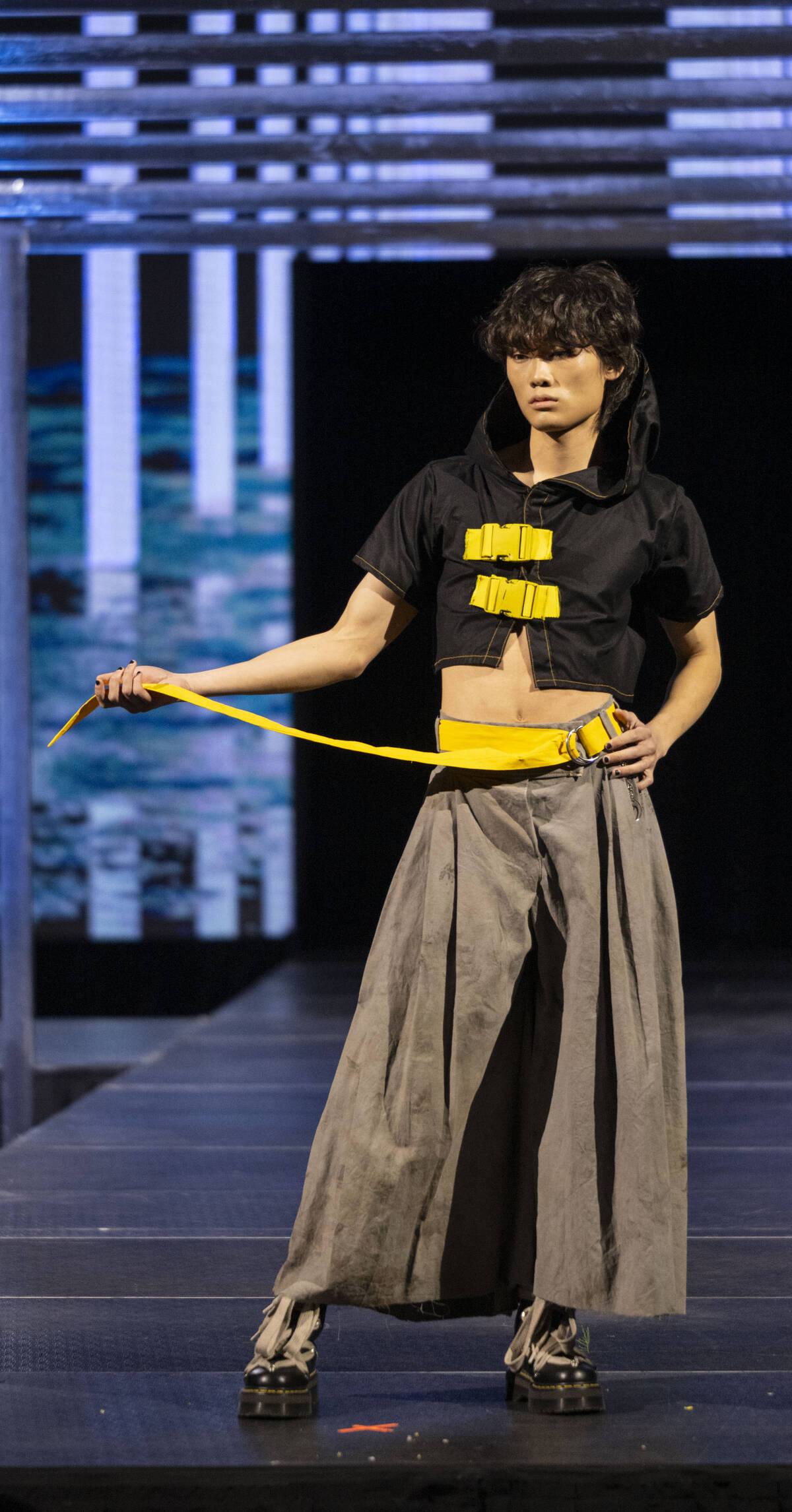 Male model wearing voluminous gray pants with a black cropped jacked and wide yellow belt and accents.