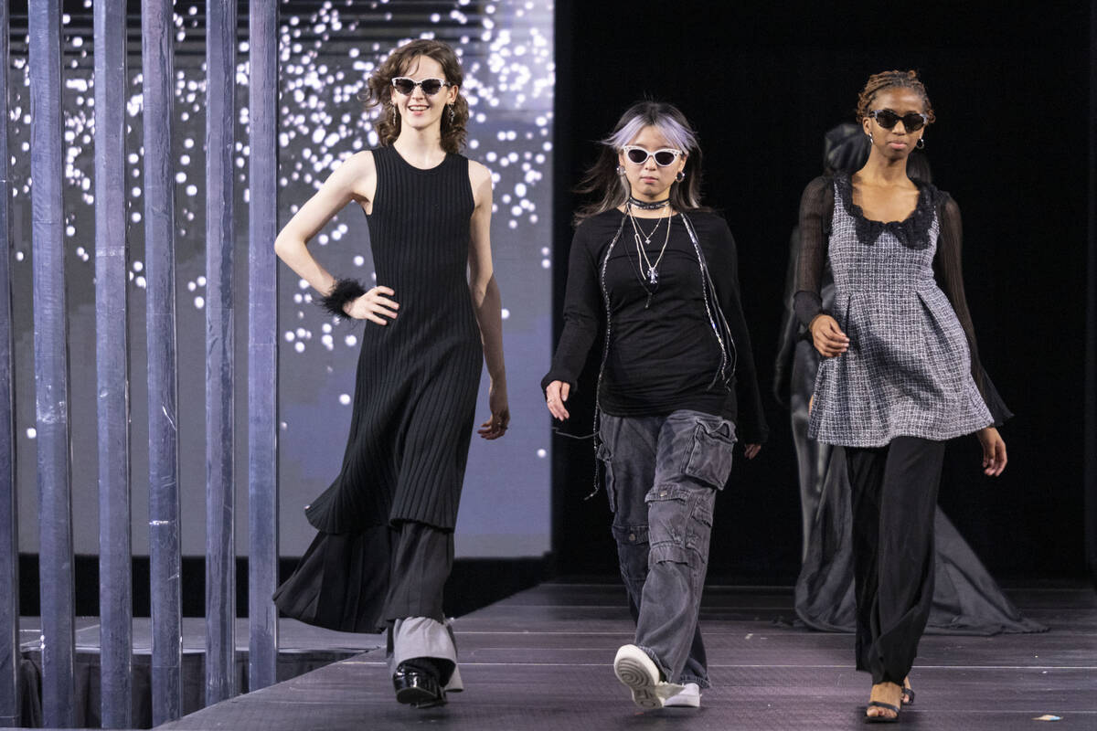 Three models wearing sleek black ensembles and sunglasses walk the runway.