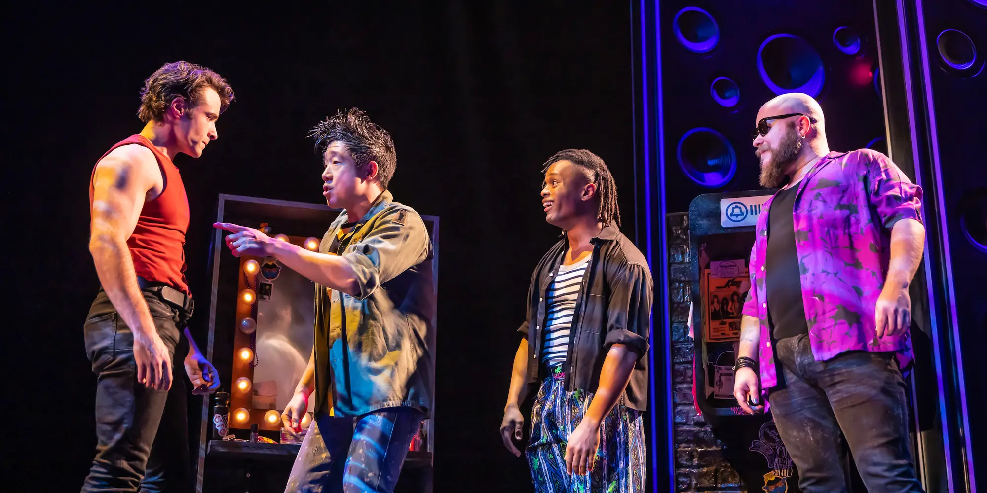On a stage, a man in a red tank top and black pants stands on the far left and listens as another man in the center of the photo points at his chest while singing and two other men to his right look on.