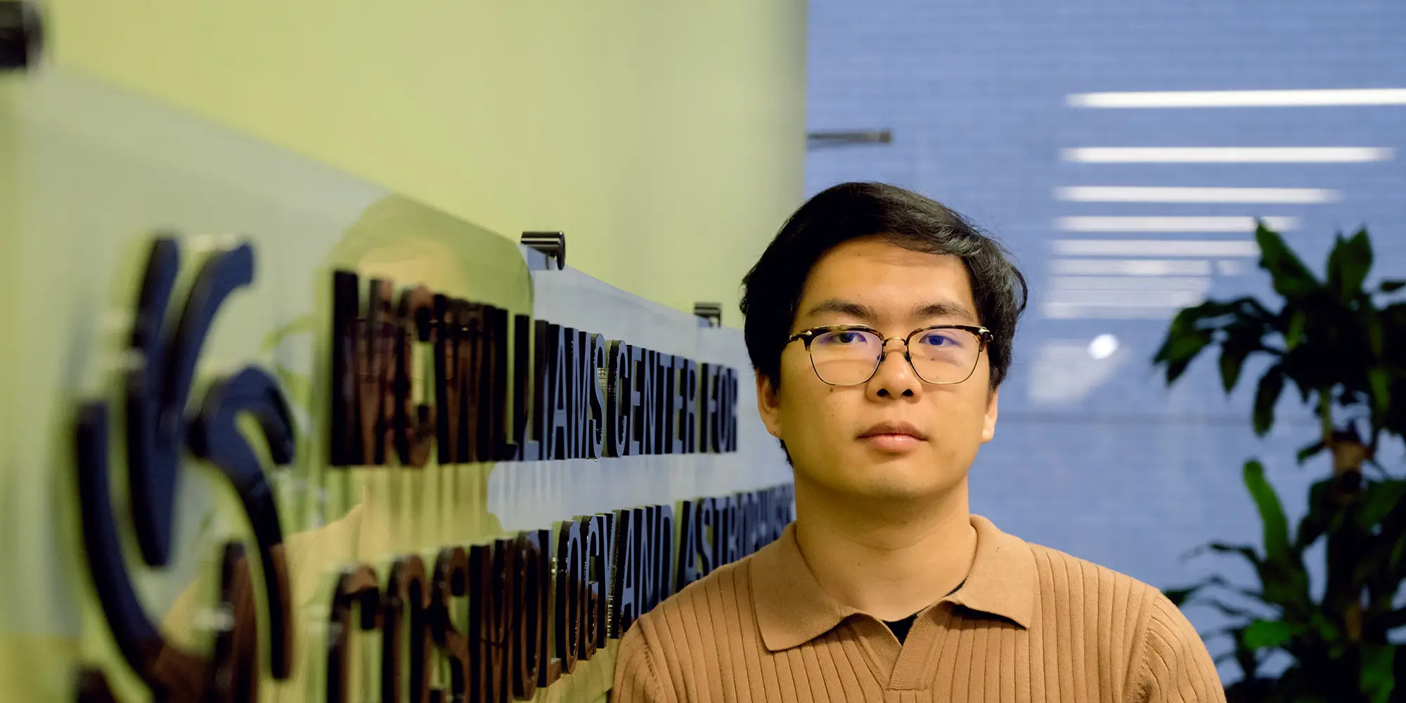 Man with glasses wearing a beige collared shirt stands near a wall with a marquee that says "McWilliams Center for Cosmology and Astrophysics" in black letters