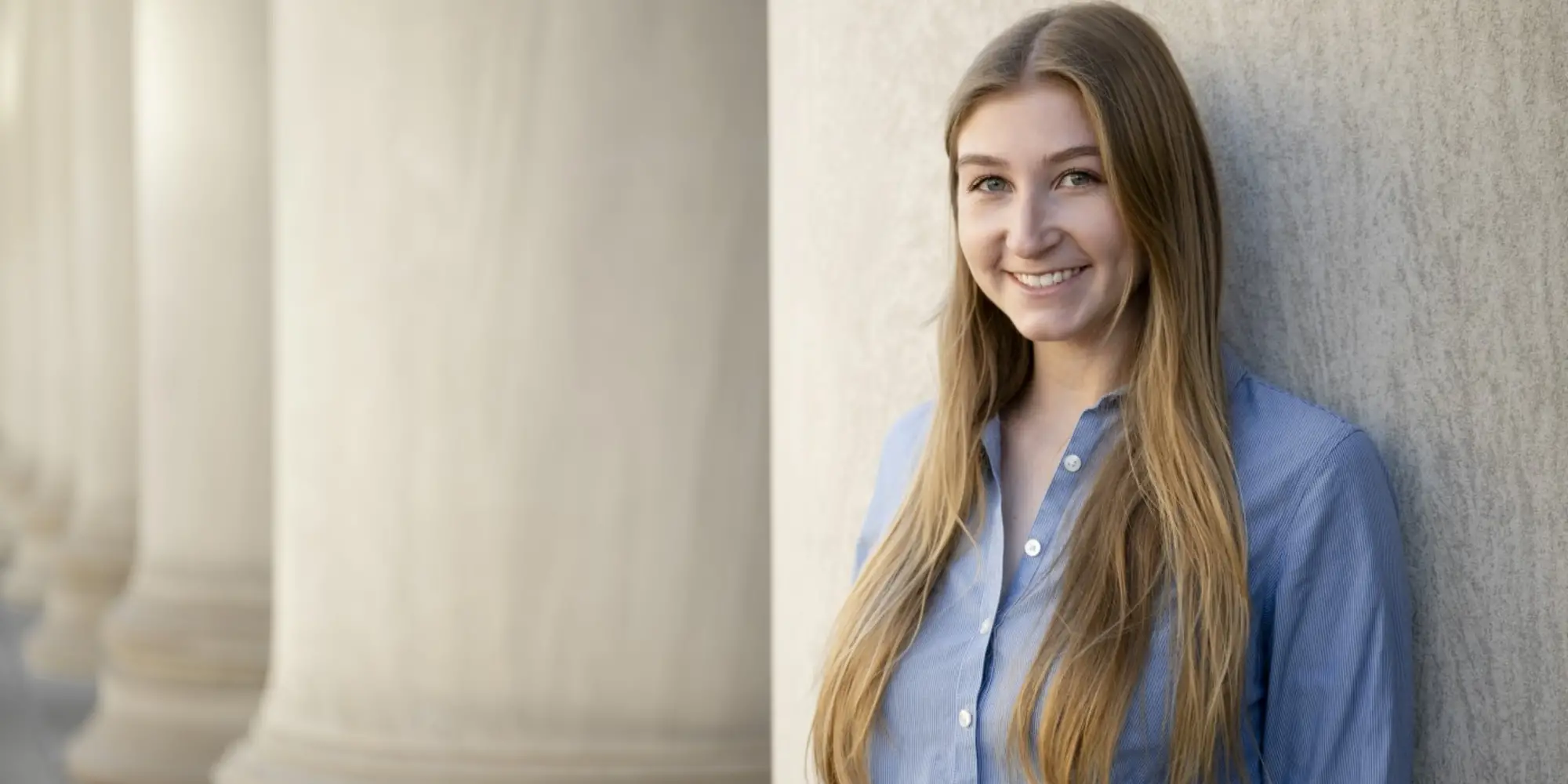 A woman wearing her long blonde hair straight down and a blue button-down dress shirt is smiling and leaning against a large stone building.