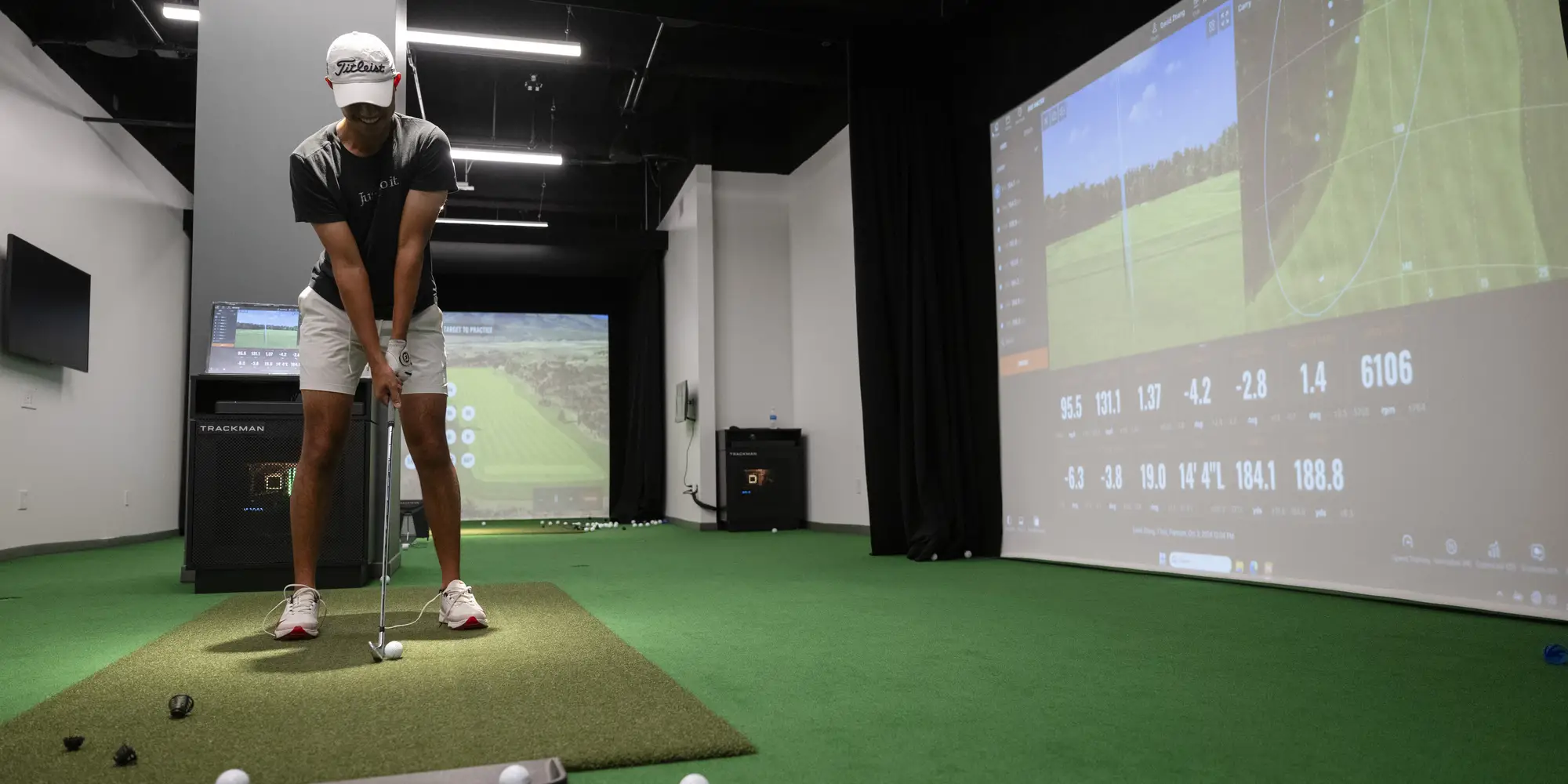 A golf team member lines up a shot holding a golf club and positioning a golf ball, about to swing and hit the ball into the screen of a simulator.
