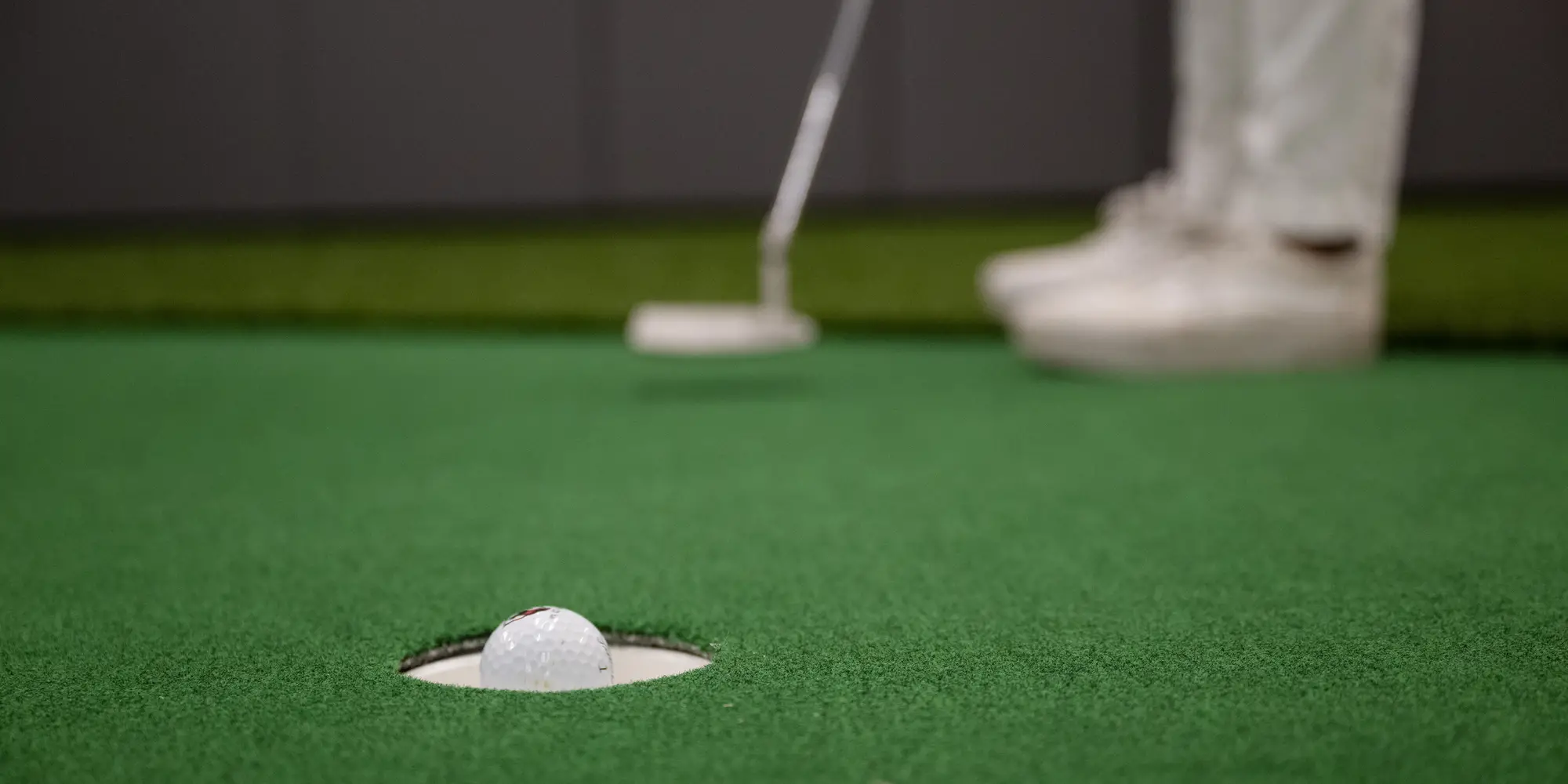 A close-up photo of a golf ball about to sink in to a putting hole shows the golfer's feet and club in the background after taking the shot.