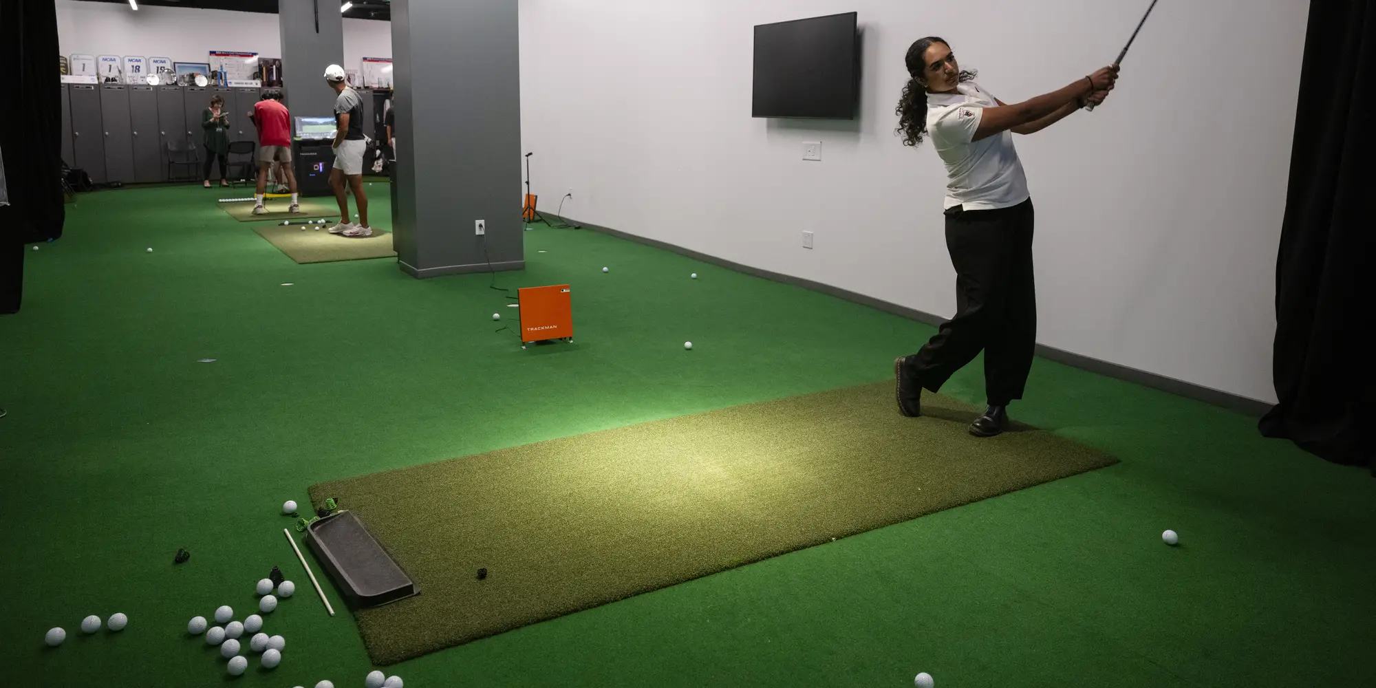 A young woman with a ponytail holds her golf club after taking a swing in a golf simulator, while other students in the background do the same in other bays of the golf teams' training room.
