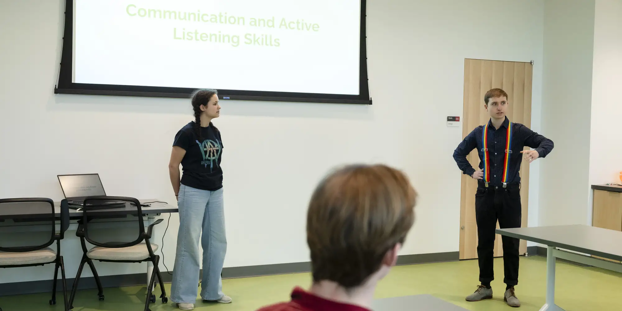 Two people stand at the front of a classroom underneath a projector screen talking while a student, listening and looking toward them, is framed in the foreground.