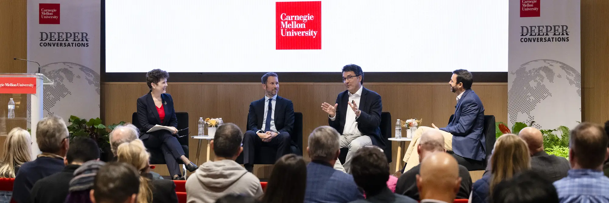 A group of four people on a stage sitting in chairs, in front of a screen that says "Deeper Conversations"