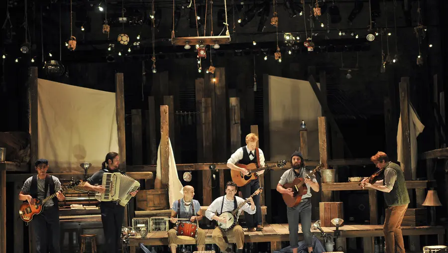 Seven men dressed as sailors on a steps of a wooden deck set on a theater stage.