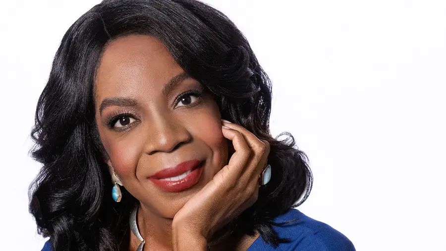 A Black woman with shoulder-length black hair in a blue dress sits in a wooden chair with her arm bent on the chair's arm and her chin resting in her hand.