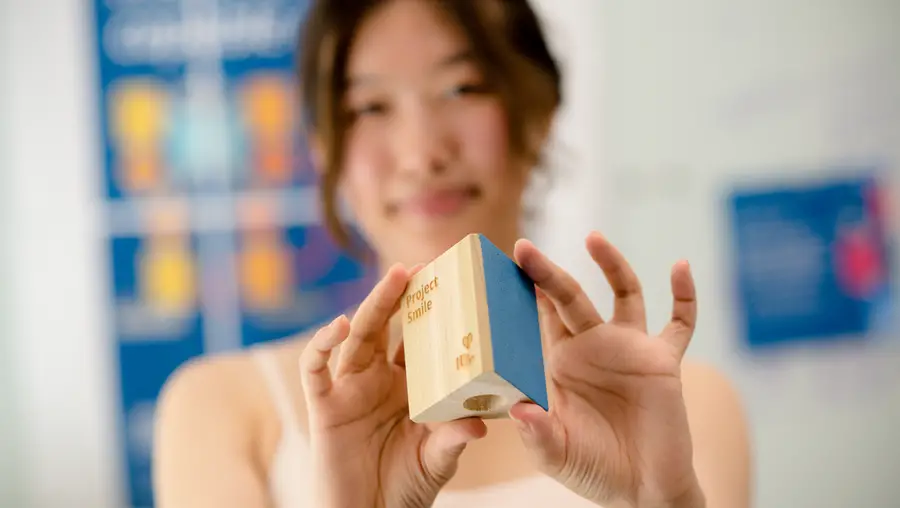 A woman out-of-focus in the background holds up a wooden block in-focus in the foreground.