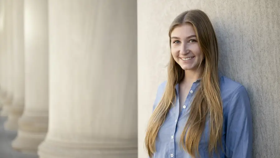 A woman wearing her long blonde hair straight down and a blue button-down dress shirt is smiling and leaning against a large stone building.