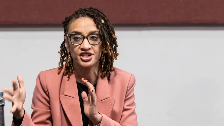 A woman with glasses wearing a salmon-colored coat gestures and talks