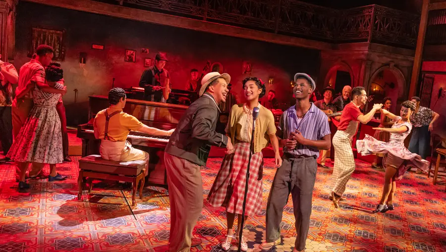 Two men and a woman stand around a microphone in the front of the photo, while behind them, in red-tinted light, other people dance and play instruments.