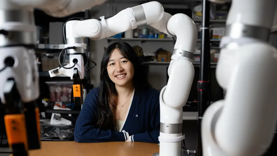 A woman with long dark hair wearing a dark blue V-neck sweater smiles at the camera while sitting behind a table with a white robotic arm attached that makes an "L" shape over the woman's head.