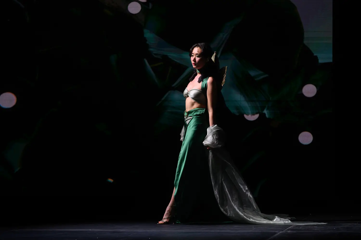 Woman wearing a pale gray bandeau top with a dramatic emerald green skirt walks mid-stride among a dark backdrop with a spotlight highlighting her profile