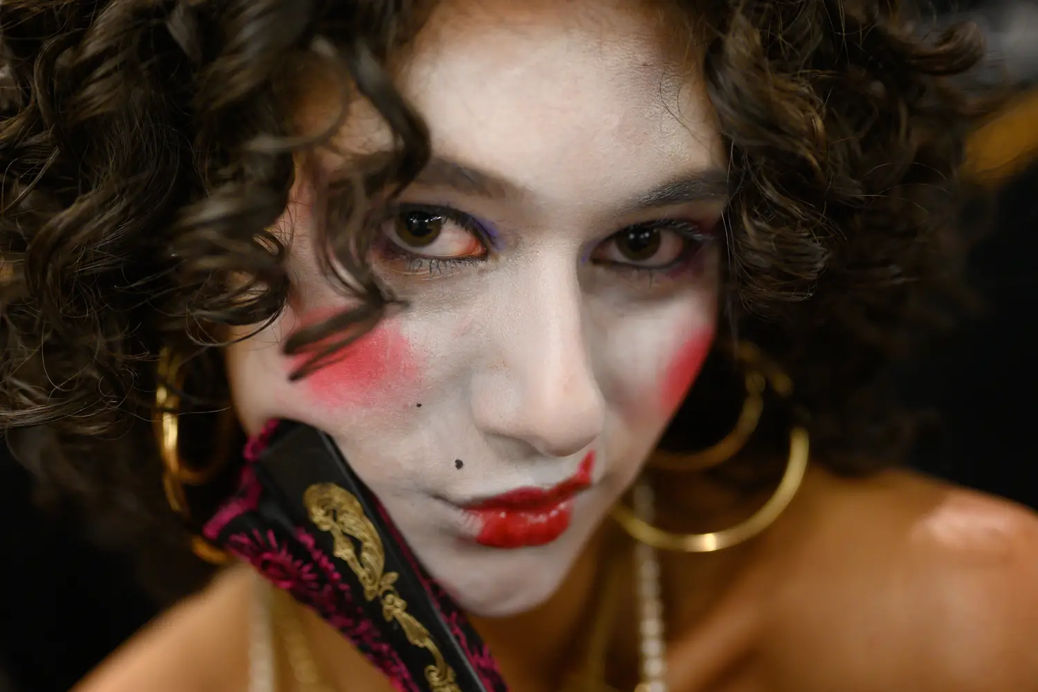 Closeup of a woman's face, wearing white face makeup with dramatic red spots of blush on her cheeks and red lipstick. She gazes into the camera, contrasting her dark hair with gold hoop earrings and a burgundy hand fan that is pressed just against her face.
