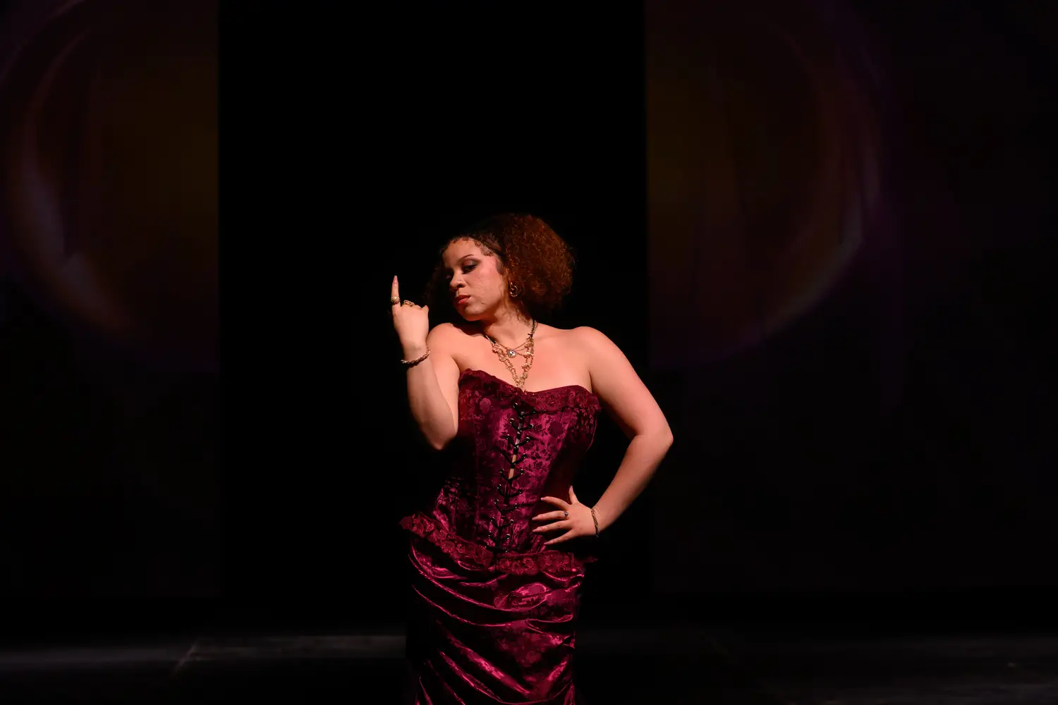 Woman poses with one hand on her hip and the other bent at the elbow pointing up. She is wearing a dramatic burgundy strapless corset-top dress with several gold necklaces layered around her neck.