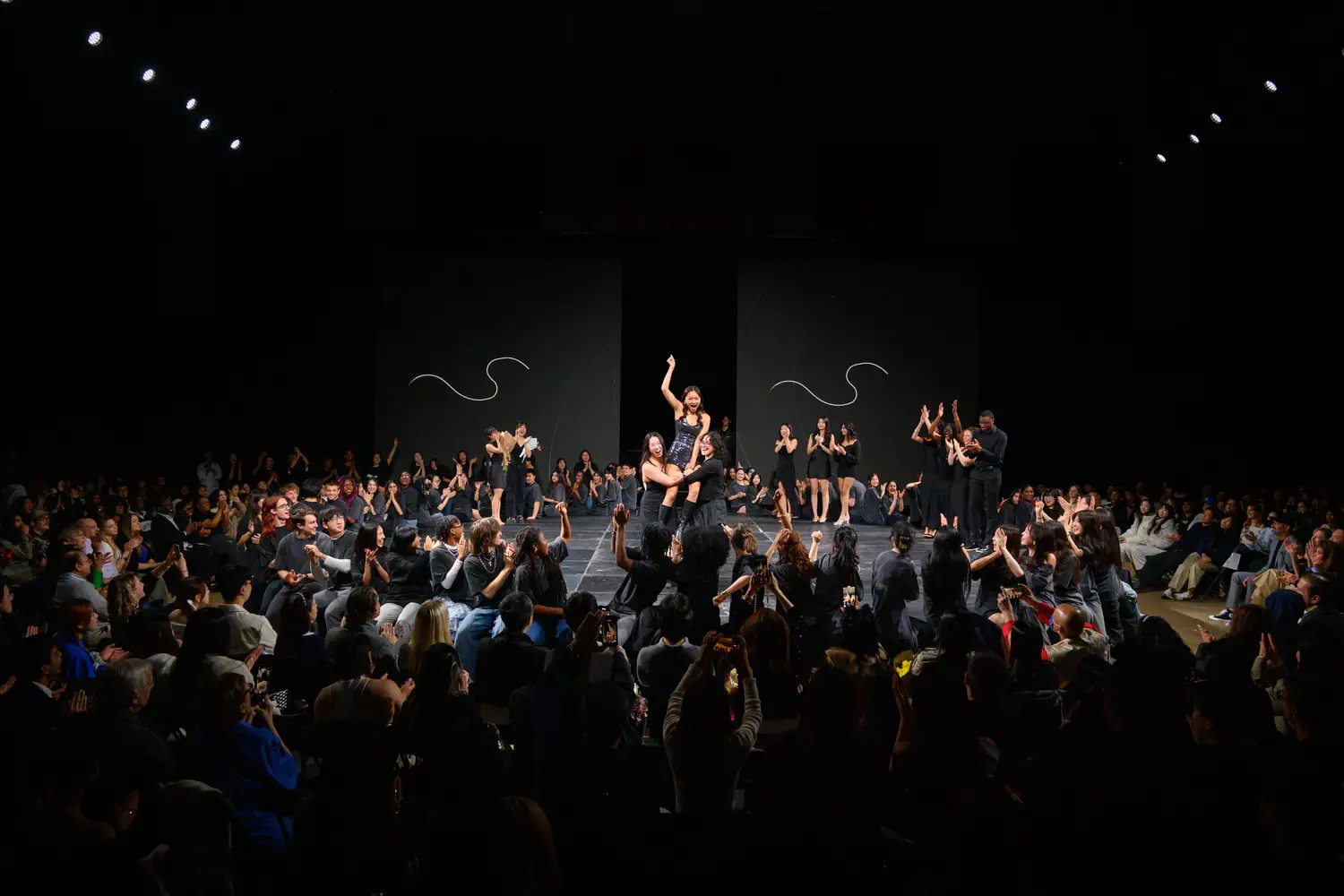 A crowd of people stands cheering and celebrating all around the circular Lunar Gala stage, with production staff and designers standing onstage and clapping. At the center of the stage is a woman waving her hand in the air while being lifted onto the shoulders of two friends; all cheering.