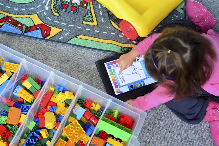 a student sits on the floor and plays with a drawing application on a tablet next to bins of colorful blocks