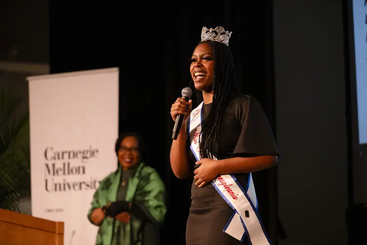 Genie Williams singing on stage wearing crown and sash with Wanda Heading-Grant standing in background