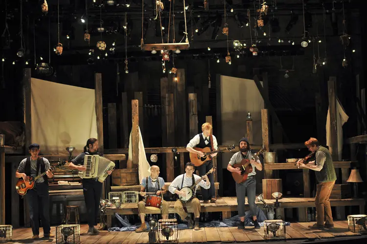 Seven men dressed as sailors on a steps of a wooden deck set on a theater stage.