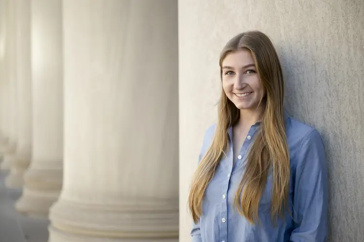 A woman wearing her long blonde hair straight down and a blue button-down dress shirt is smiling and leaning against a large stone building.