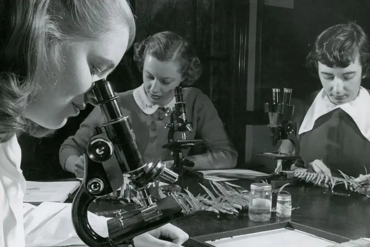A black and white historic photo of women using a microscope. 