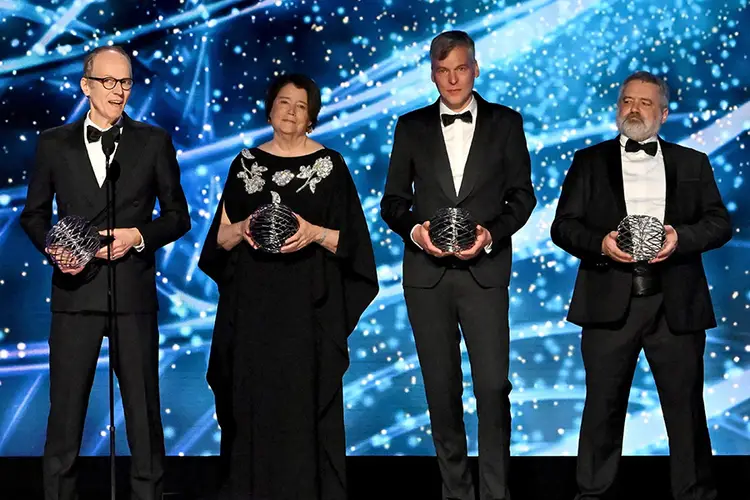 Three men and one woman hold spherical wire sculptures on a stage. The background is blue and filled with flashes of light.